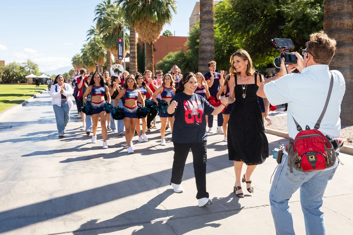 A group of cheerleaders and a marching band walking down a pathway lined with palm trees. In the foreground, two women are holding hands and walking, with one wearing a black dress and the other a dark shirt with red and white graphics. Several cheerleaders, dressed in matching blue and red uniforms with pom-poms, are behind them, along with members of a marching band carrying instruments like sousaphones and trumpets.