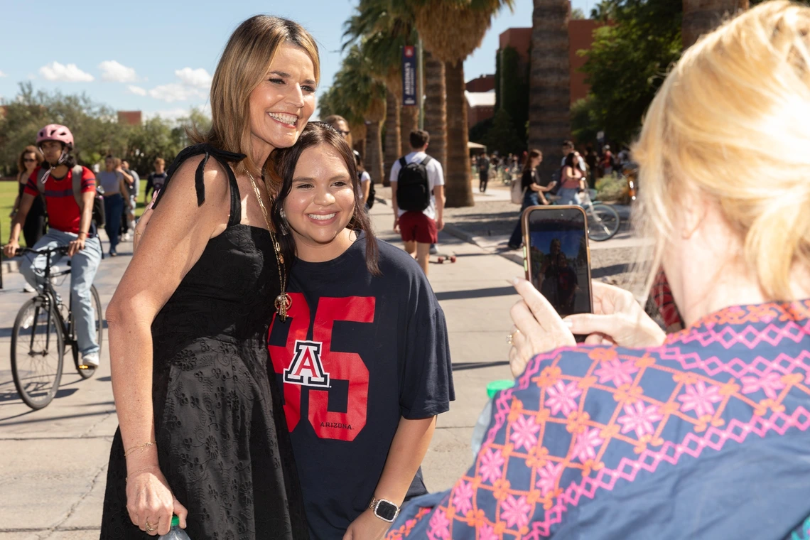 Two women are posing for a photograph on a sunny day. The woman on the left is wearing a black dress and has long hair. She is smiling broadly as she stands next to a young woman wearing a navy-blue T-shirt featuring a large red number "95" with the letter "A" and the word "ARIZONA" underneath. The young woman also smiles at the camera. A third person in the foreground, with blonde hair, is taking a photo with a smartphone.