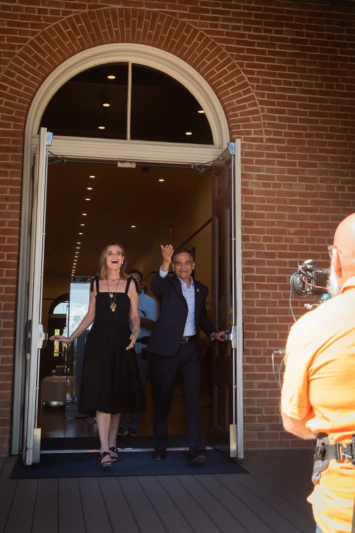 A man and a woman exiting a building through open double doors. The doorway is set in a red brick wall, topped with a semicircular window above the doors. The woman on the left is wearing a black dress and sandals, and she appears to be speaking or laughing. The man on the right is in a suit, gesturing outwardly with his hand.