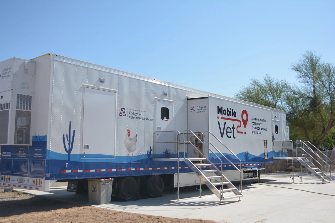 A large mobile veterinary clinic trailer against a clear blue sky. The trailer is white with blue accents and features images of animals, including a chicken and a dog. The left side displays the logo and text for the University of Arizona College of Veterinary Medicine. Metal stairs with railings lead to entrances on the side of the trailer. Various graphics, including cacti, are painted along the bottom.