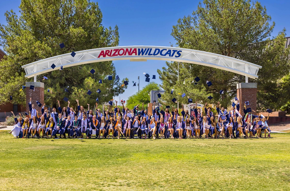 Several college students throw their caps into the air under an archway with the words Arizona Wildcats on it.