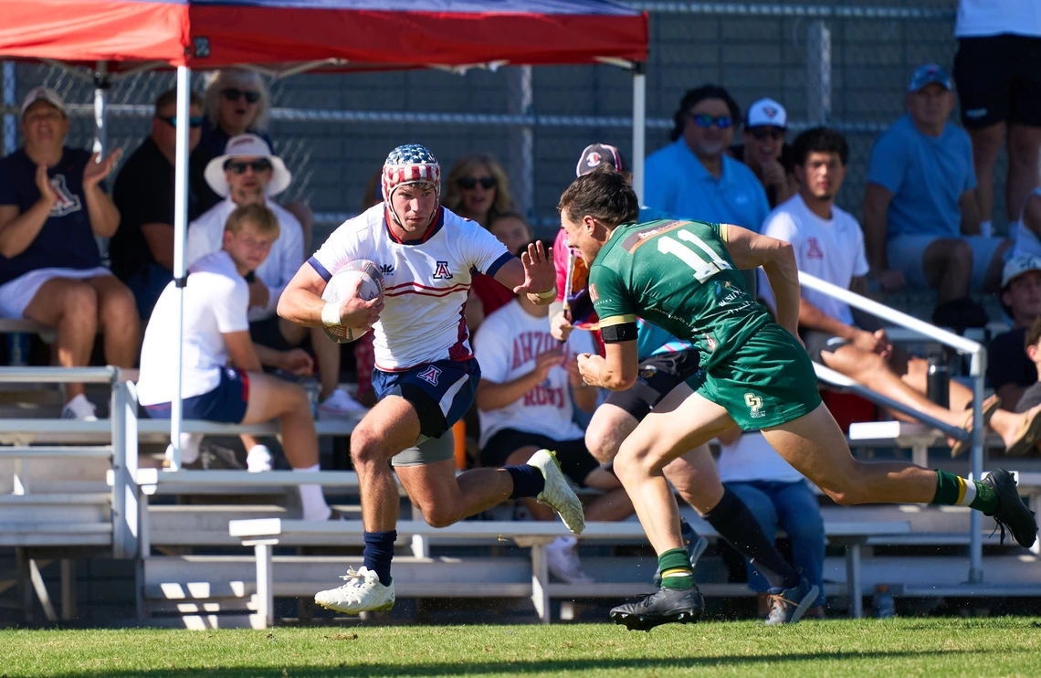 A rugby match with two male players in action. The player on the left, in a white jersey with blue and red accents, holds a rugby ball and is sprinting forward. He wears a protective headband with a star pattern. The player on the right, wearing a green jersey marked with the number 11, is approaching to tackle. Both players are on a grassy field with sunlight casting shadows.