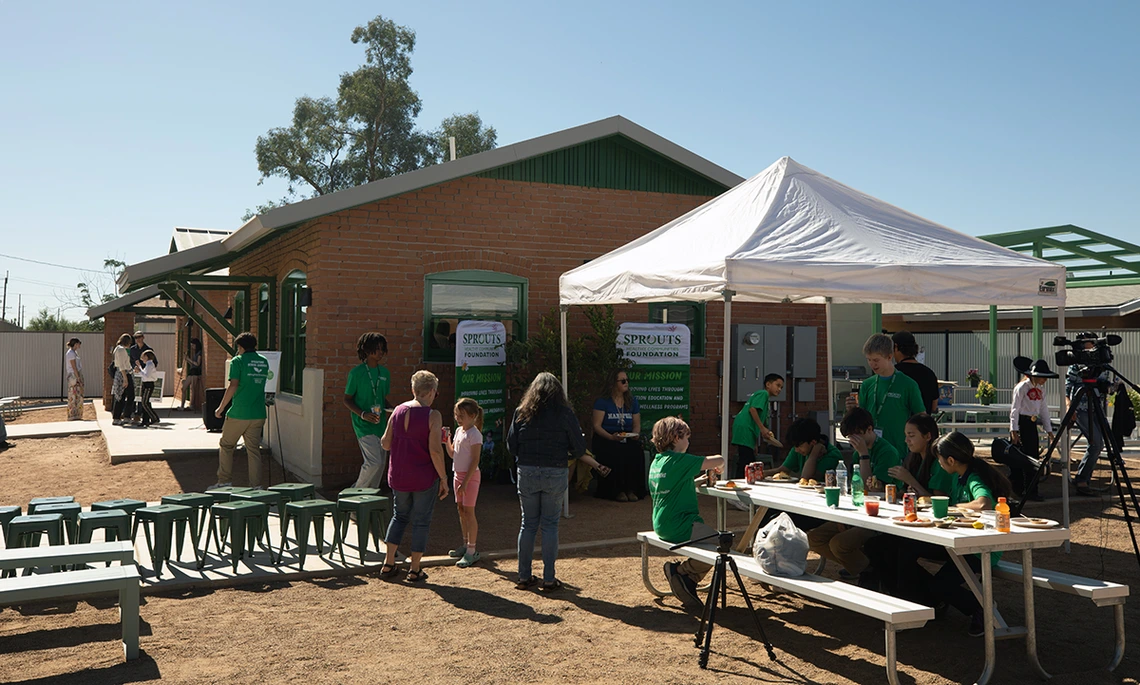 People gather outside the newly renovated Sprouts House during a sunny daytime event. A white tent shades a table where participants in green shirts sit and eat, while others talk nearby. The restored brick building with green trim stands in the background with banners showing the Sprouts Foundation’s mission.