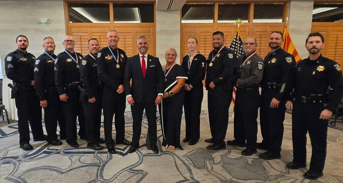 A group of University of Arizona Police Department officers stand in a line with U.S. Rep. Juan Ciscomani at the Veteran Servant Leader Award ceremony. The officers wear formal uniforms and medals, and U.S. and Arizona flags are displayed behind them.