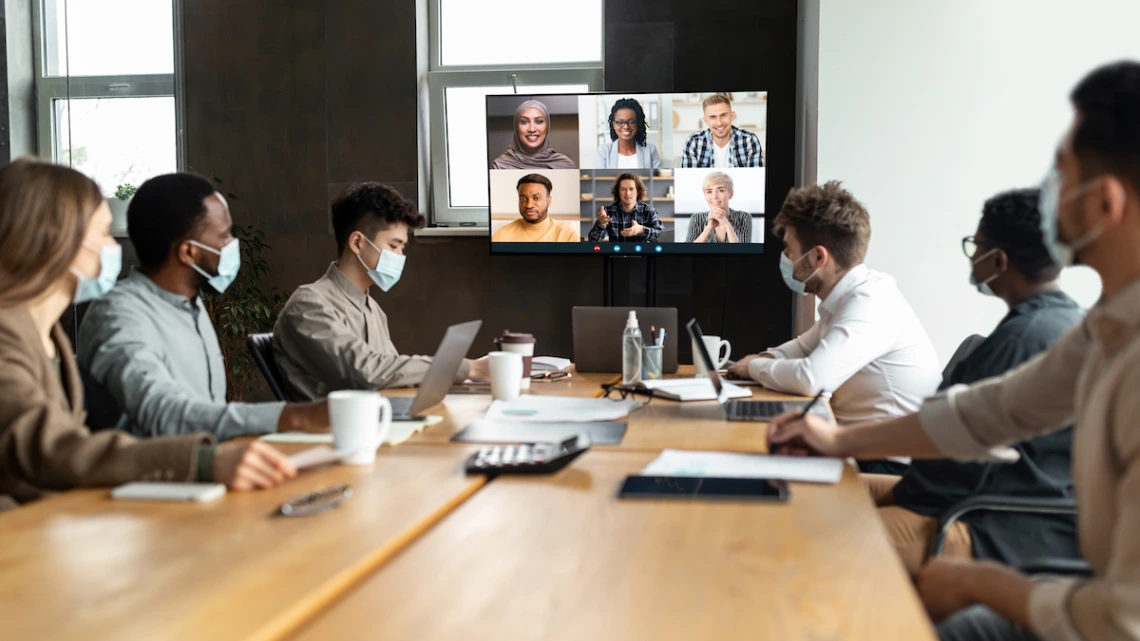 a group of people at a table looking at a screen displaying virtual meeting participants Hybrid meetings can be a challenge, leaving virtual attendees feeling ignored. But they can be successful with some planning and flexibility to make sure all participants have equal opportunity to contribute.