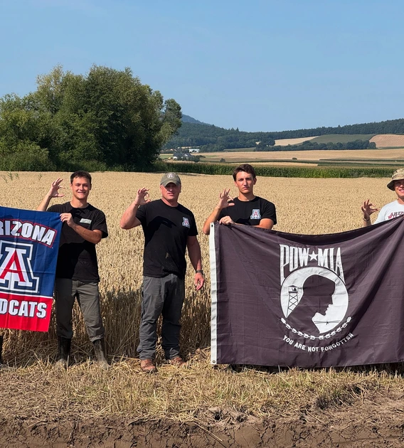 Five people stand in a golden wheat field holding two flags—one red, white, and blue Arizona Wildcats flag and one black POW*MIA flag with the words “You Are Not Forgotten.” They wear casual outdoor clothing, some with boots and hats, and make the University of Arizona “Wildcat” hand sign against a backdrop of rolling farmland and blue sky.