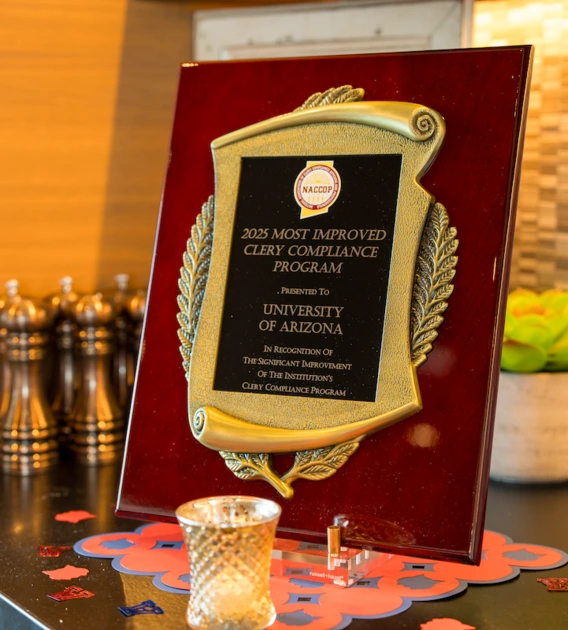 A close-up of a plaque reading "2025 Most Improved Clery Compliance Program, presented to University of Arizona," displayed on a countertop with small decorative items around it.