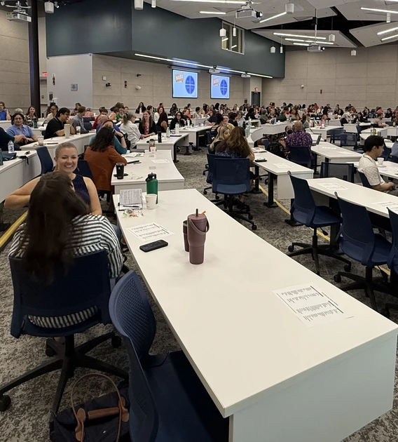 Attendees fill a large lecture hall during the July 30 Conflict Resolution and De-escalation Skills Symposium at the University of Arizona. Participants sit in groups at long white tables, engaging in discussion during an interactive portion of the event. Multiple presentation screens display a slide with the word “practice” and a blue circular graphic divided into four sections.