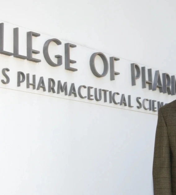 Jack R. Cole, wearing a brown plaid suit and patterned tie, stands smiling in front of a white wall with large lettering that reads "College of Pharmacy Skaggs Pharmaceutical Sciences."