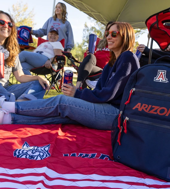 People relax under a canopy at a tailgate on a sunny day, sitting on a red University of Arizona blanket. A navy backpack with “Arizona” and the “A” logo is in the foreground.