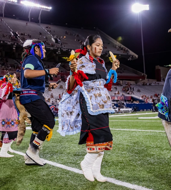 Hopi dancers performing a sacred corn dance in traditional tribal clothing on a football field with stadium lights shining down
