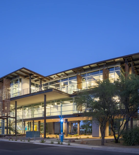 Modern three-story building with large glass windows, lit from inside at dusk, surrounded by trees and landscaping, with a blue emergency call box in front.