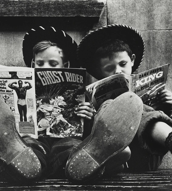 Two boys in cowboy hats sit side by side with their feet stretched out toward the camera, each reading a comic book. One holds The Ghost Rider while the other reads a Zane Grey western. The image is in black and white, highlighting their worn shoes and absorbed expressions.