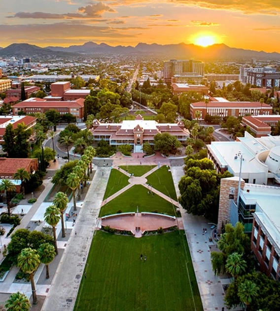 Aerial view of the University of Arizona campus at sunset, with Old Main at the center, the grassy Mall stretching toward the mountains in the distance, and the sky glowing with orange and purple clouds.
