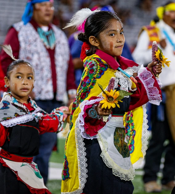 Two young children in traditional Hopi regalia perform a dance on the field, holding corn and flowers, during the Hopi Recognition Football Game at Arizona Stadium, with adult participants standing behind them.