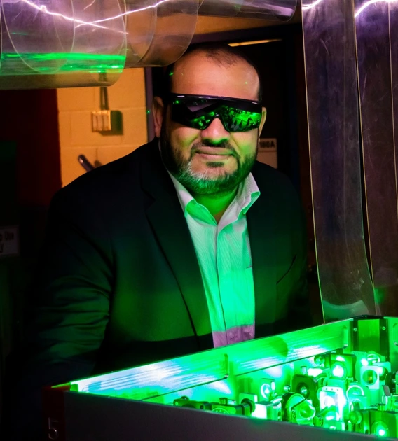A man wearing safety glasses and a suit observes a green-lit optical experiment with lasers and mirrors in a laboratory setting.