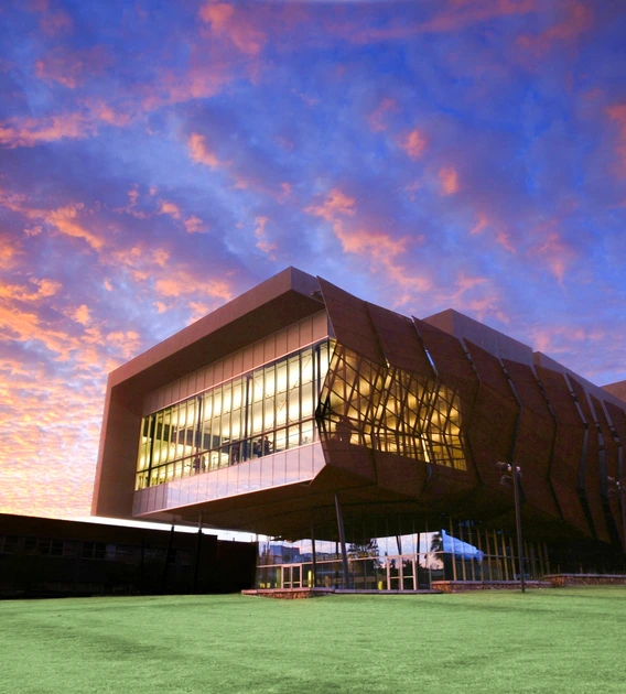 A modern, angular building with a geometric design situated against a dramatic sky during sunset. The structure features large glass windows framed by brown, asymmetric panels. The upper section of the building extends outward, creating an overhang supported by columns. The sky behind the building is a vibrant mix of orange and purple hues, with scattered clouds reflecting the sunset colors.