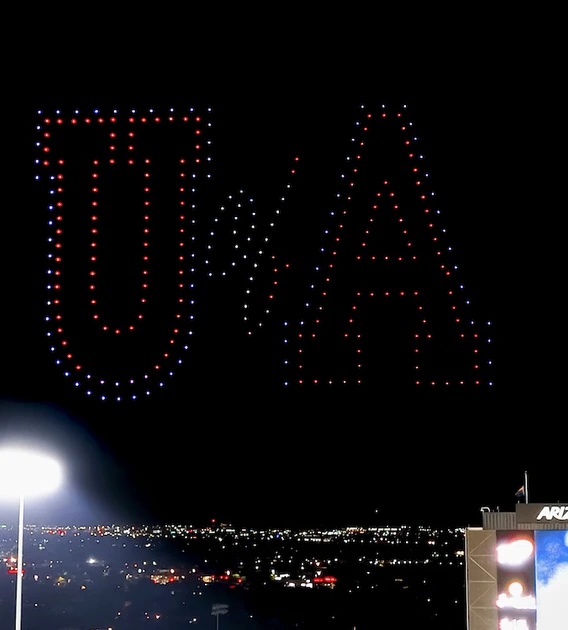A nighttime drone show above Arizona Stadium forms a red, white, and blue "U of A" in the sky during a University of Arizona football game. The illuminated letters appear over the lit stands and scoreboard as the city lights of Tucson glow in the distance.
