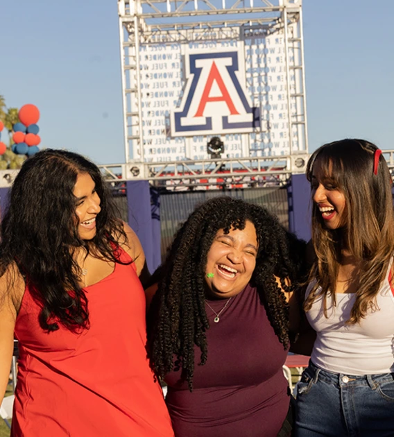 Three women smile and laugh in front of the Unviersity of Arizona logo.