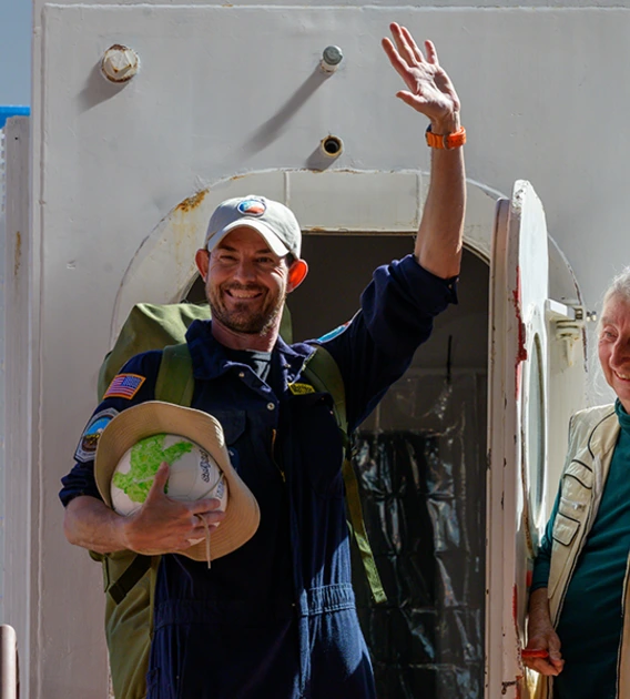 A person opens the door to a sealed habitat as a second person exits, waving and smiling.