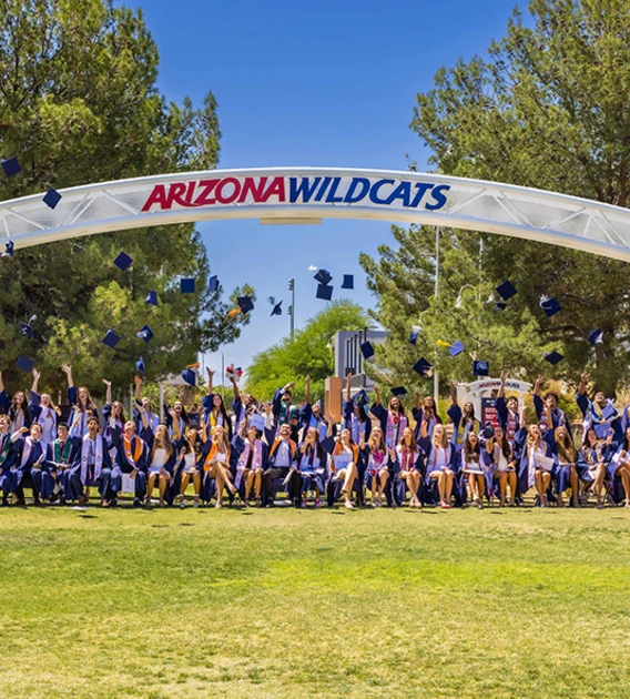 Several college students throw their caps into the air under an archway with the words Arizona Wildcats on it.