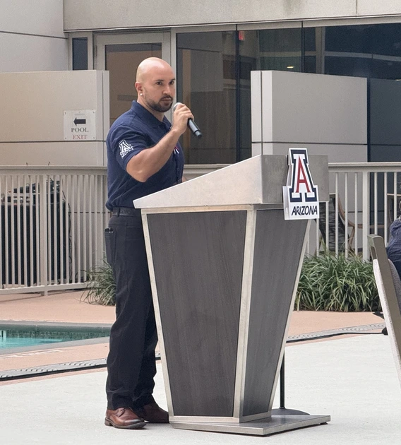 A man in a navy blue shirt speaking into a microphone at a podium. The podium is positioned near a swimming pool and features the University of Arizona logo with the word "ARIZONA" on it. In the background, there is a railing and plants adjacent to a modern building with large windows.
