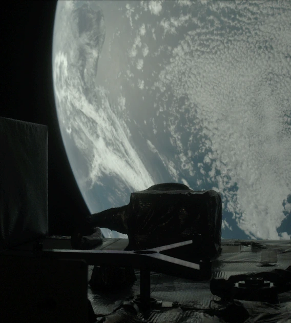 Earth fills most of the image, a moody blue globe mostly covered with tiny blueish white clouds like carpet pile or sheep’s wool. In the foreground, the instrument panel is dark enough to be nearly in silhouette, like a city skyline. A tall thin rectangle juts up from the left side of the panel, like a tall building, which is part of the SamCam. In the center, a shorter, squatter polygon shape is the OLA instrument. A slightly lighter colored Y shape lays on its side in front of the OLA.