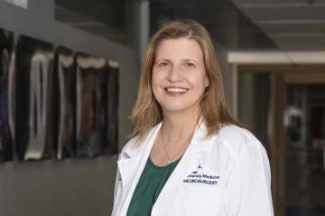 A woman wearing a white medical coat with “Neurosurgery” and “Banner – University Medicine” embroidered on it stands smiling in a modern hallway, suggesting a clinical or academic hospital setting. She has straight, shoulder-length hair and wears a dark green blouse under her coat.