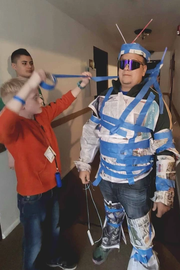 Alejandro Vazquez standing in a room while children assemble a costume on him using a roll of tape