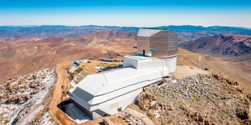 Rubin Observatory from above on its desert summit site, with a vast expanse of desert mountains receding into the background. Rubin is a boot-like shape at center, with a long white service building extending toward us and to the left and angular silver dome rising on the right.