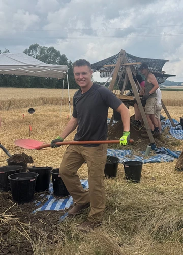 A man in a dark T-shirt and khaki pants smiles while holding a red shovel in a field, standing near black buckets on a blue-striped tarp. Behind him, other people work under a mesh canopy, sifting soil through large wooden screens as part of an outdoor excavation or fieldwork project.