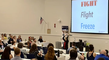 Catherine Tornbom, a facilitator for the July 30 Conflict Resolution and De-escalation Skills Symposium, stands at the front of a lecture hall addressing attendees. She holds a black object above her head while a large screen behind her displays the words “FIGHT,” “Flight,” and “Freeze” in red, gray, and blue text. About two dozen faculty, staff, and administrators sit at desks, listening attentively. An American flag hangs on the wall, and an Office of Public Safety banner is visible near the screen.