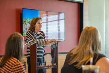 A woman speaks at a clear podium with “Arizona Sands Club” on the front, addressing an audience. Behind her, a screen displays the words “Clery Compliance Committee Members” along with a list of names.