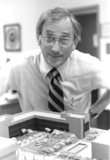 Black-and-white photo of Jack R. Cole in a dress shirt, tie and glasses, leaning over a scale model of a building.
