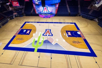 An overhead view of the basketball court at McKale Center, featuring a large red, white, and blue block “A” at center court with a stylized desert design including a green saguaro cactus, blue mountains, and an orange sun. The court is bordered in blue with “Arizona” marked on each baseline and Big 12 Conference logos inside both key areas. A large scoreboard hangs above the court.