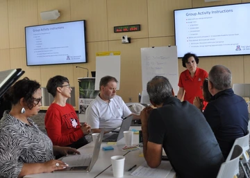  A group of six people sit around a table in a meeting room at the University of Arizona, listening to a woman in a red U of A polo shirt who is speaking to them. Laptops, papers, and coffee cups are on the table, and two screens behind them display “Group Activity Instructions.”