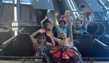 Five people, including three children, smile and raise their hands while riding the Star Wars: Rise of the Resistance attraction at a Disney park. Stormtrooper figures and sci-fi set pieces are visible in the background.
