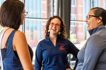 Three women smile and talk inside a bright campus building. The woman in the center wears red glasses, University of Arizona earrings, and a navy pullover with a red-and-blue “A” logo.