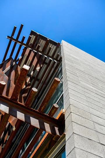 The exterior of a masonry building, next to which run a series of rusted steel beams.