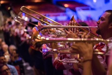 Close-up of trumpet players performing indoors, with brass instruments gleaming under stage lights and an audience visible in the background.