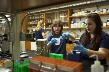 Two students wearing gloves conduct lab work at Biosphere 2, measuring and preparing samples at a bench surrounded by scientific equipment, while another researcher works in the background.