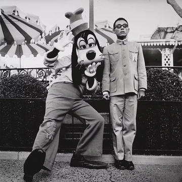 Black-and-white photograph of a man in a buttoned uniform standing stiffly and expressionless beside a costumed Goofy character, who is crouched in a playful pose with one hand raised and the other on his chin. Amusement park umbrellas and landscaping are visible in the background.