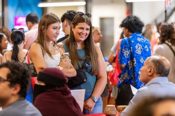 Two women smile while talking to a man seated at a table during the University of Arizona’s Destination Arizona event. Other attendees are visible in the busy background.