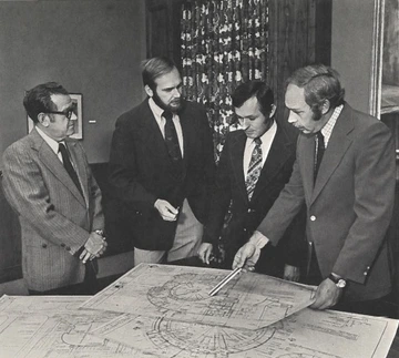 A black-and-white photograph showing four men in suits standing around a large table with architectural blueprints spread across it. The man on the right is gesturing towards the blueprints with a pen, indicating some details to the others. The men appear to be engaged in a discussion, examining the plans closely.