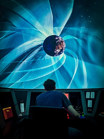 A person seated at a control desk, facing a large, curved screen. The screen displays a digital, artistic visualization of Earth, surrounded by blue, swirling auroras or abstract lines, depicted in shades of blue and white.