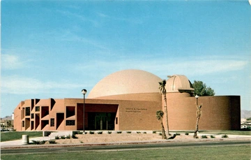 The exterior of a large brick planetarium under a clear blue sky. The building features a prominent dome-shaped structure at the center and a smaller dome with a slit aperture to the right. The front facade is decorated with geometric patterns and consists of vertical ridges with alternating rectangular recesses.
