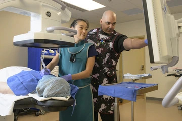Two medical professionals in protective lead aprons stand beside a patient during a procedure, with one person preparing a syringe and the other pointing to a monitor while guiding the process.