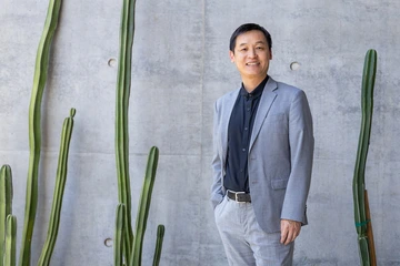 A person wearing a light gray suit and black shirt stands smiling beside tall cacti against a concrete wall.