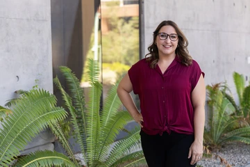 A person wearing a burgundy blouse and black pants stands smiling with one hand on their hip in front of a modern concrete wall and green ferns.