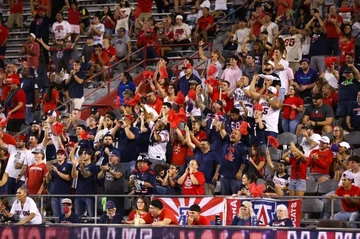 A large crowd of University of Arizona football fans cheer in the stadium stands, many wearing red, blue, and white clothing and waving red pom-poms. Some fans clap, shout, or hold up the “Wildcat” hand sign as they celebrate during a game.